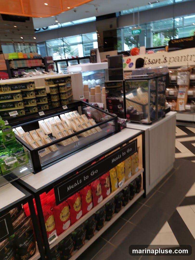 A view inside a modern convenience store, showcasing various food items and a checkout area. In the foreground, shelves are stocked with packaged snacks and pre-made meals labeled "Meals to Go." A refrigerated display case holds sandwiches. In the background, more shelves display colorful products, and a sign reads "Madison's Pantry." An "EXIT" sign is visible above glass doors leading outside.