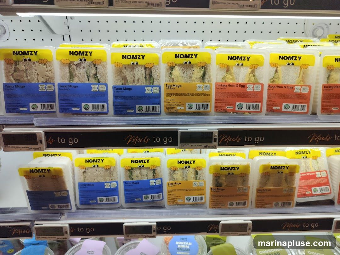 A refrigerated display case filled with pre-packaged sandwiches from the brand NOMZY. The sandwiches are arranged on two shelves. The top shelf features Tuna Mayo, Egg Mayo, and Turkey Ham & Egg sandwiches. The bottom shelf also shows Tuna Mayo and Egg Mayo sandwiches, along with other items not fully visible. Each sandwich is in clear plastic packaging with a yellow label featuring the NOMZY logo and cartoon eyes. Price tags are visible below some of the items.