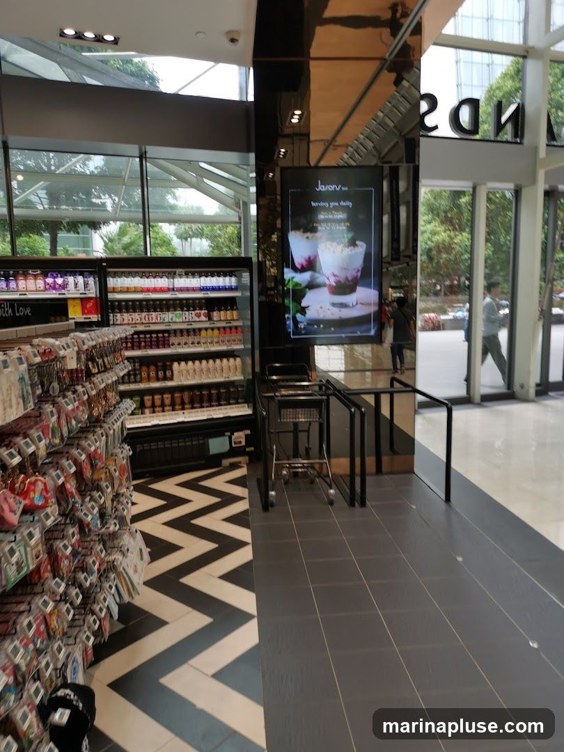 The image shows the interior of a store with shelves stocked with various products, including drinks and small merchandise. A digital display screen shows an advertisement for "Jason's" with a picture of a dessert. A shopping cart is visible near the display. The floor has a distinctive black and white chevron pattern in one section and grey tiles in another. Large windows allow natural light to enter, and reflections of the exterior and interior are visible on a mirrored surface.