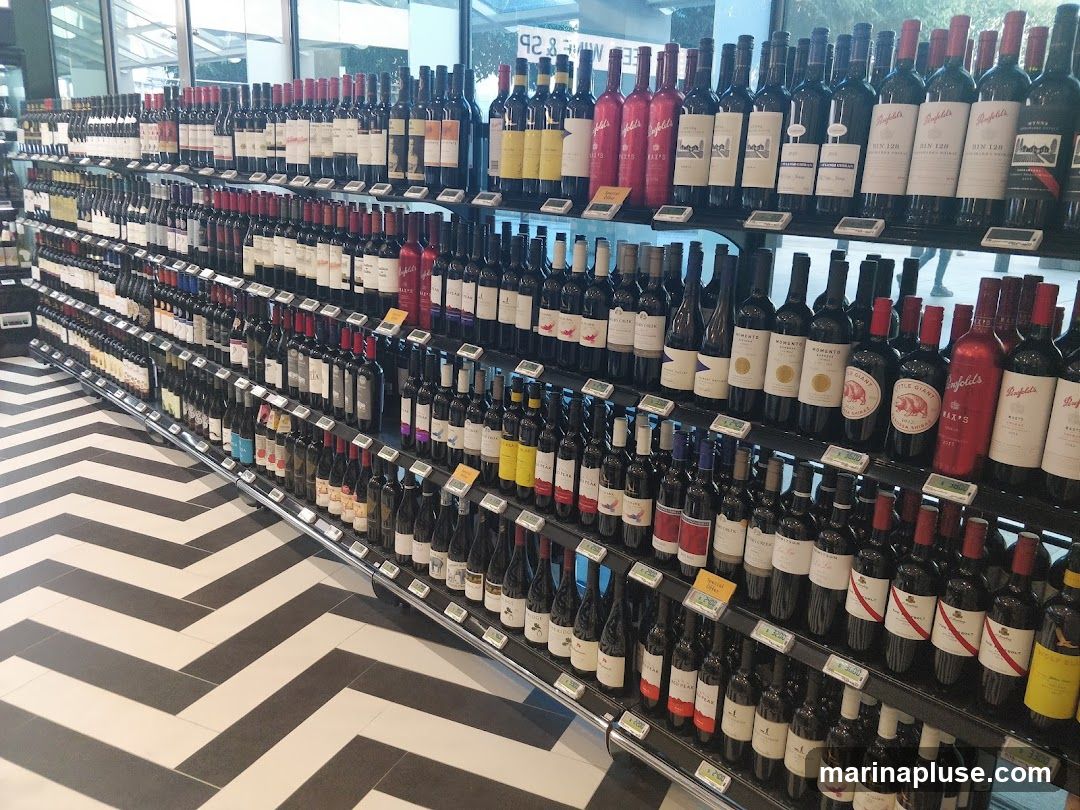 A wide selection of wine bottles are displayed on shelves in a store. The bottles are arranged by type and brand, with some bottles standing upright and others lying on their sides. The shelves are well-lit, and the bottles are clearly visible. The floor has a black and white chevron pattern.