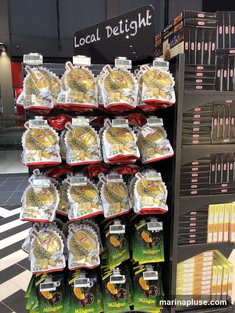 A display of packaged snacks, primarily durian and mango flavored, with prices shown on digital tags. The sign above reads "Local Delight".
