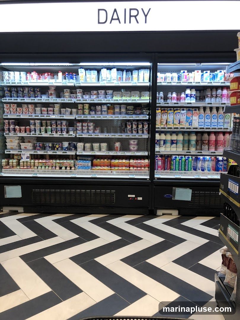 A refrigerated display case filled with various dairy products, including yogurts, milk, and other beverages. The aisle has a distinctive black and white chevron-patterned floor. The word "DAIRY" is displayed in large white letters above the refrigerated units.