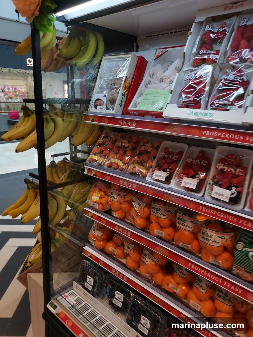 A refrigerated display case filled with various fruits, including bananas, strawberries, tangerines, and grapes. The fruits are packaged in plastic containers and boxes, with some featuring Japanese text and branding. The display is well-lit, highlighting the freshness of the produce.