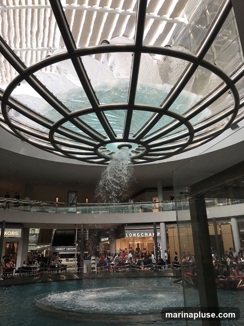 An indoor view of a large shopping mall featuring a central water fountain with water cascading from a circular structure above. Surrounding the fountain are various shops, including Michael Kors and Longchamp, with people seated at tables near the water. The mall has multiple levels with balconies overlooking the fountain area.