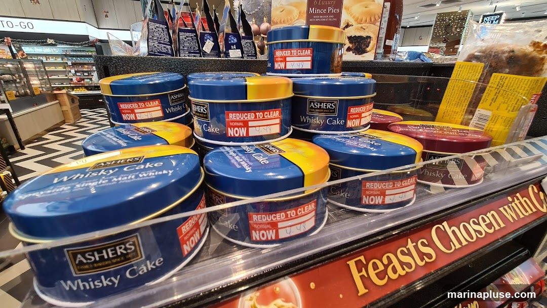 A close-up view of several round tins of Ashers Whisky Cake stacked on a store shelf. Each tin is blue and yellow or blue and red, with the Ashers logo and "Whisky Cake" prominently displayed. Red "Reduced to Clear" stickers with a price of 4.48 are affixed to many of the tins. In the background, a refrigerated display case with food items and other products can be seen, along with signage for "6 Luxury Mince Pies" and "Feasts Chosen with C". The floor has a black and white chevron pattern.