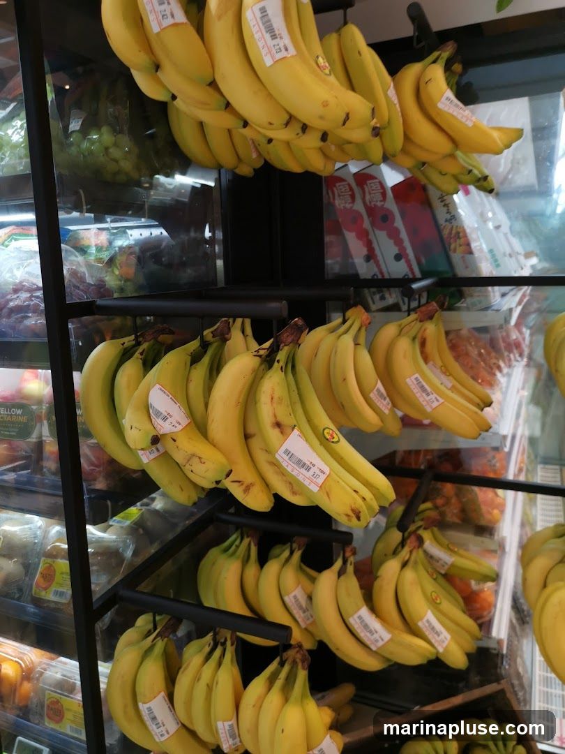 A close-up shot of several bunches of ripe yellow bananas hanging from black metal racks in a grocery store. Some bananas have price tags attached. In the background, other produce like grapes and oranges are visible, along with packaged goods on shelves.