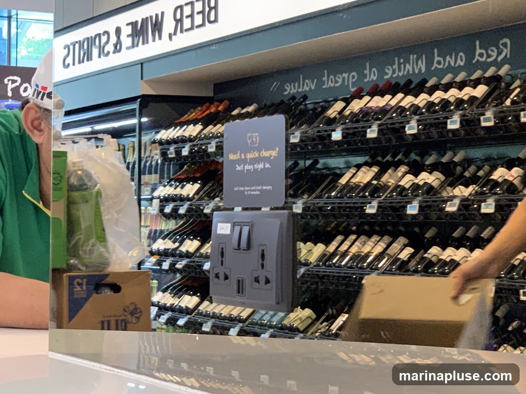 A display of wine bottles on shelves in a store. A sign above reads "BEER WINE & SPIRITS" and "Red and White Wine of Great Value". A charging station is visible on a shelf. A person in a green shirt and white hat is partially visible on the left.