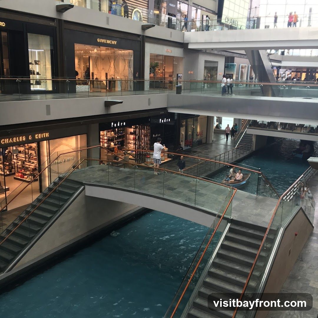 An interior view of a multi-level shopping mall featuring an indoor canal with a boat carrying passengers. Bridges connect different sections of the mall, spanning over the blue water. Various retail stores, including Givenchy and Charles & Keith, are visible on the upper levels, with their storefronts displaying merchandise. People are seen walking on the bridges and walkways, and some are browsing in the shops.