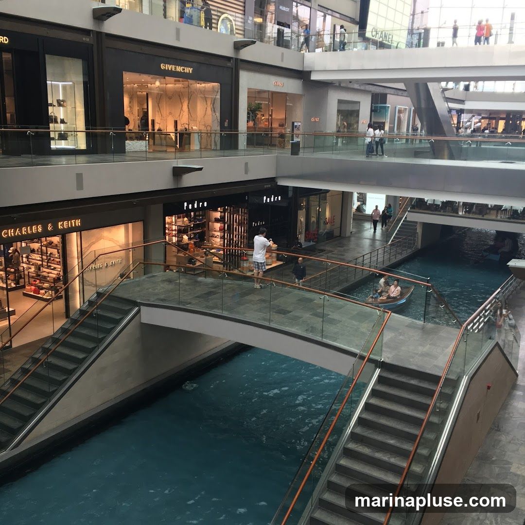An interior view of a multi-level shopping mall featuring an indoor canal with a boat carrying passengers. Bridges connect different sections of the mall, spanning over the blue water. Various retail stores, including Givenchy and Charles & Keith, are visible on the upper levels, with their storefronts displaying merchandise. People are seen walking on the bridges and walkways, and some are browsing in the shops.