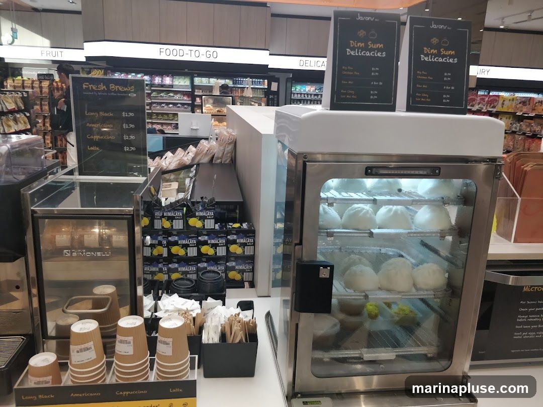A food and drink counter with various items on display. In the foreground, there is a coffee machine and cups for different beverages like Long Black, Americano, Cappuccino, and Latte. To the right, a glass display case shows steamed buns, likely dim sum. Behind these, shelves are stocked with products, and overhead signs indicate 'FRUIT', 'FOOD-TO-GO', 'DELICAT', and 'IRY'. Two menu boards for 'Dim Sum Delicacies' are also visible.