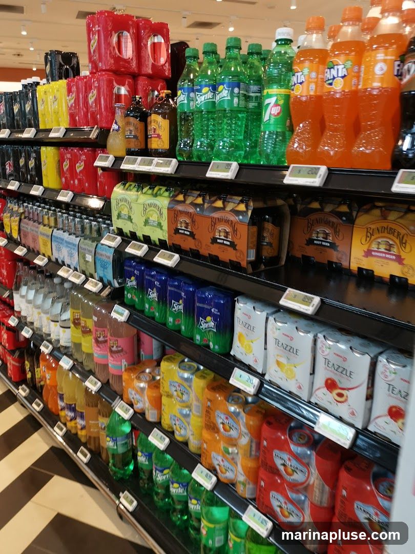 A wide selection of bottled and canned beverages are displayed on shelves in a store. The products include various brands of soda, juice, and other drinks, with prominent displays of Sprite, Fanta, 7UP, Bundaberg Root Beer, and San Pellegrino.
