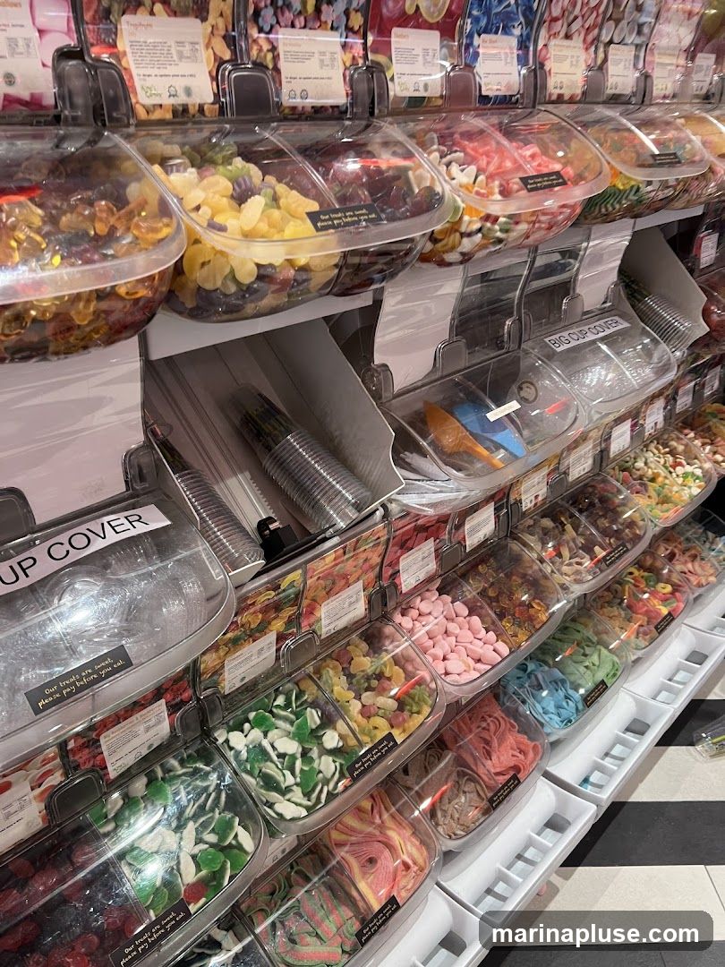 A wall display of various candies in clear bins, with scoops and cups available for customers. The bins contain a wide assortment of gummy candies, sour candies, and other sweets in different colors and shapes. Some bins have small labels with text, including "Our treats are sweet, please pay before you eat." and "BIG CUP COVER".