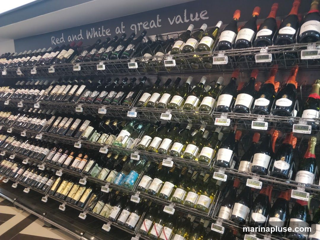 A wall of shelves filled with various bottles of wine, predominantly red and white. The bottles are arranged in rows and sections, with some featuring prominent labels and corks. A sign above the display reads "Red and White at great value." Price tags are visible on some of the shelves.