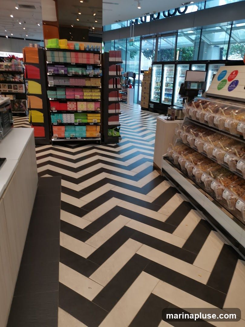 The image shows the interior of a store with a distinctive black and white chevron pattern on the floor. To the left, shelves are stocked with colorful boxed items. In the background, refrigerators are visible, and to the right, a display case holds bulk food items. The overall impression is a modern and clean retail environment.