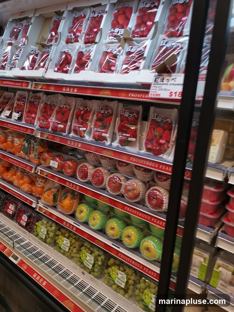 A refrigerated display case filled with various fruits, including strawberries, oranges, apples, and grapes. The strawberries are packaged in clear plastic containers, some with Japanese labels. The other fruits are also packaged for sale. Red and gold decorative trim with Chinese characters runs along the shelves.