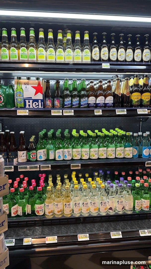 A refrigerated display case filled with various bottled beverages, including beer, cider, and soju. The top shelf features bottles of Somersby Apple and Pear cider, followed by bottles of Kopparberg Pear Cider and Thatchers Gold Cider. Below that are bottles of Heineken beer and other assorted beers. The middle shelf is dominated by numerous green bottles of Jinro soju in flavors like Chamisul, Green Grape, and Plum. The bottom shelf shows more bottles of soju and other assorted drinks with colorful caps. Price tags are visible on some shelves.