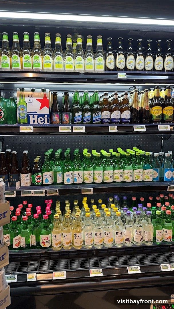 A refrigerated display case filled with various bottled beverages, including beer, cider, and soju. The top shelf features bottles of Somersby Apple and Pear cider, followed by bottles of Kopparberg Pear Cider and Thatchers Gold Cider. Below that are bottles of Heineken beer and other assorted beers. The middle shelf is dominated by numerous green bottles of Jinro soju in flavors like Chamisul, Green Grape, and Plum. The bottom shelf shows more bottles of soju and other assorted drinks with colorful caps. Price tags are visible on some shelves.