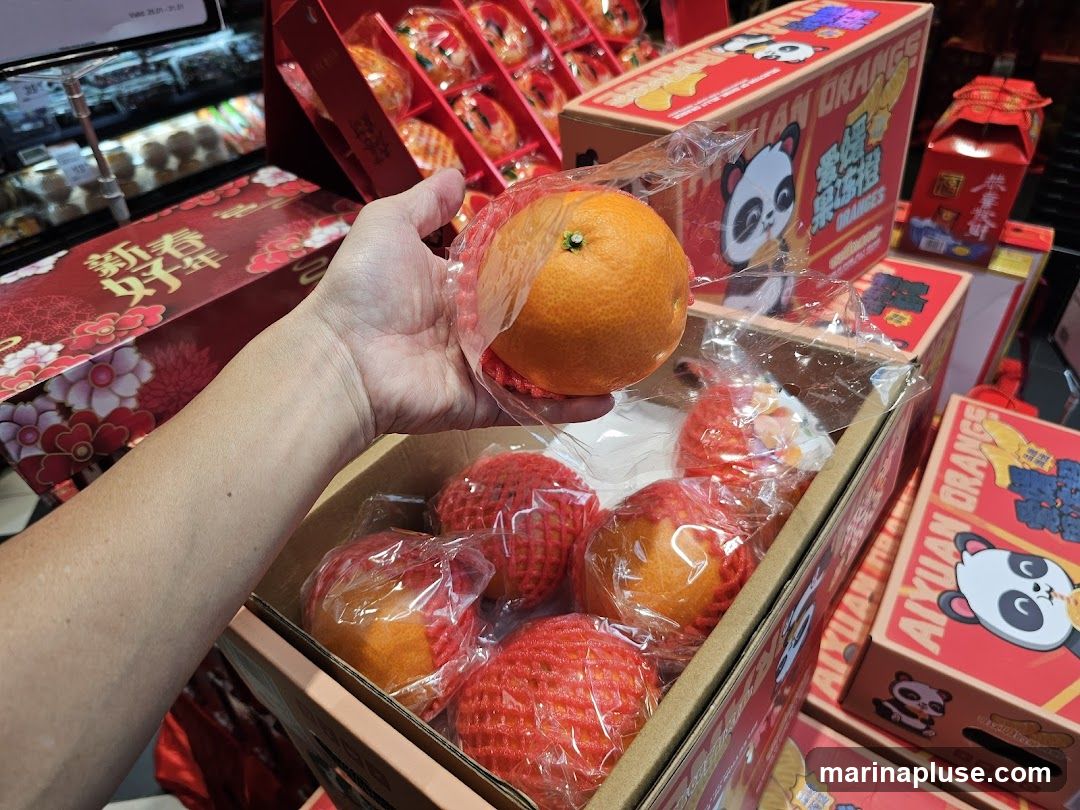 A hand holds an orange over a box of oranges. The oranges are individually wrapped in red netting and clear plastic. The boxes are red and decorated with Chinese characters and cartoon pandas. Some boxes are labeled "AIYUAN ORANGE". In the background, more gift boxes and refrigerated shelves are visible.