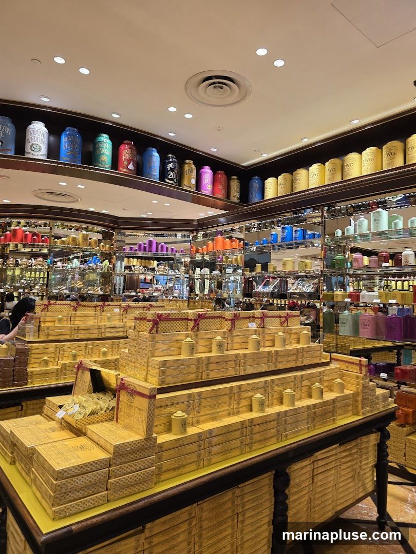 A brightly lit interior of a tea shop, showcasing an elaborate display of tea boxes and canisters. The foreground features a tiered pyramid of yellow boxes, some open to reveal tea bags. The shelves in the background are filled with a vast array of colorful tea canisters, organized by color and brand. A person is visible on the left, browsing the merchandise.