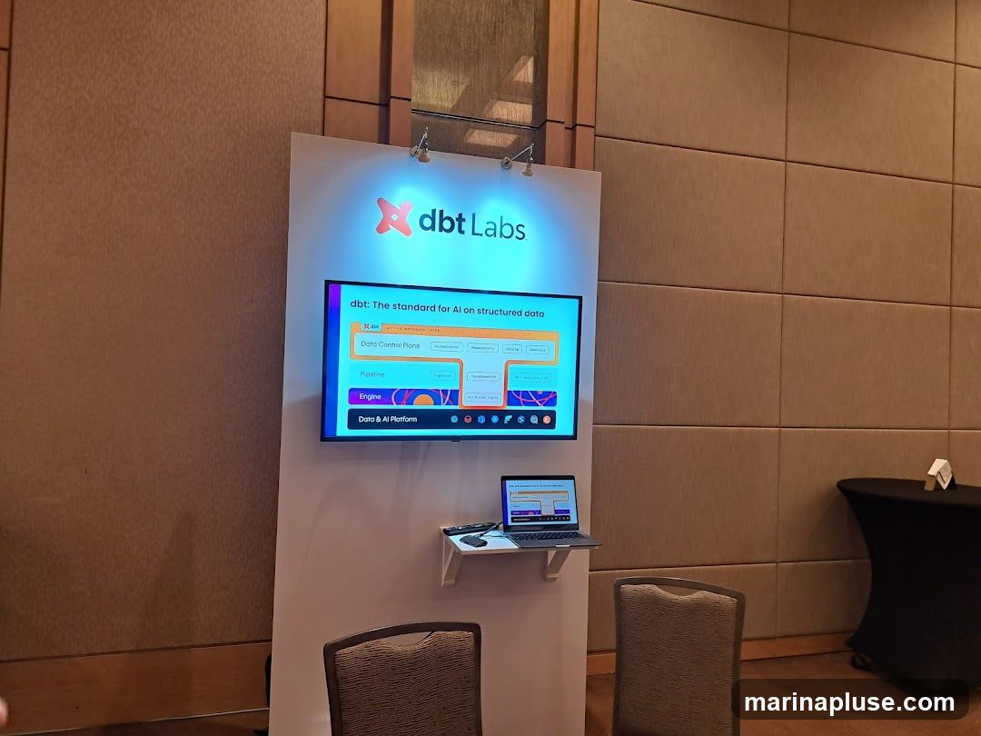 A display featuring the dbt Labs logo and the tagline "dbt: The standard for AI on structured data." A screen shows a diagram with sections for "Data Control Plane," "Pipeline," "Engine," and "Data & AI Platform." A laptop is placed on a shelf below the screen. Two chairs are visible in the foreground.