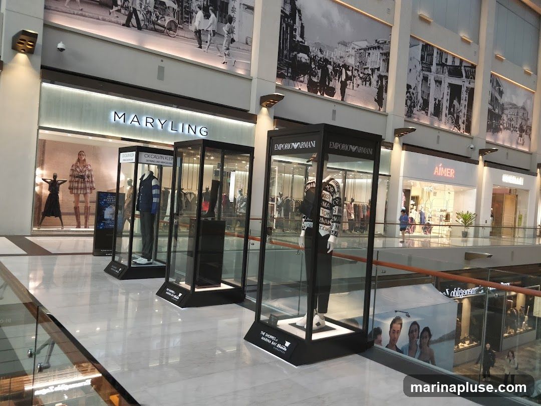 A view of a modern shopping mall interior with multiple storefronts visible. In the foreground, there are several glass display cases showcasing clothing and mannequins from brands like Emporio Armani and CK Calvin Klein. Behind these displays, storefronts for MARYLING, AIMER, and other retailers are visible. Large black and white murals depicting historical street scenes adorn the upper walls of the mall.