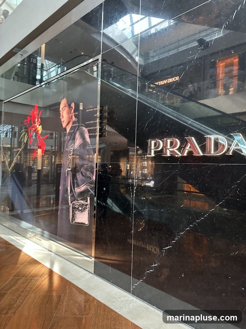 A large advertisement featuring a man in a coat and a small handbag is displayed on the glass facade of a Prada store. The Prada logo is prominently visible on the black marble exterior. Reflections of escalators and other store interiors are visible in the glass.