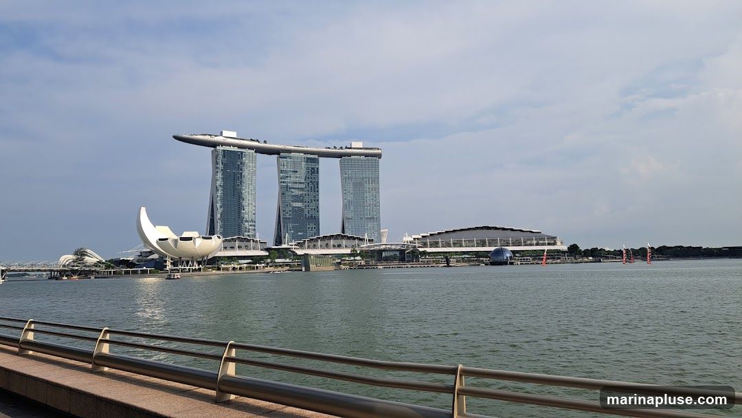A wide shot of the Marina Bay Sands complex in Singapore, with its iconic three towers and the ArtScience Museum. The image is taken from across the water, with a railing in the foreground. Several sailboats are visible in the distance. The sky is partly cloudy.