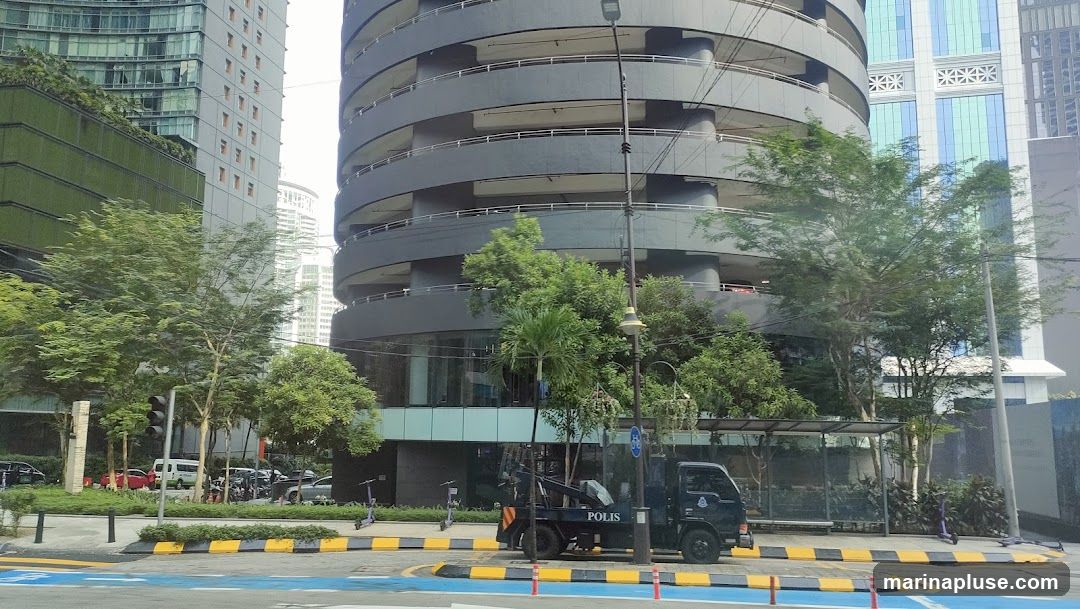 A view of a street in a city, featuring several modern buildings with glass facades and a distinctive spiral parking garage. Trees and greenery are interspersed throughout the scene. A dark-colored truck with the word "POLIS" visible on its side is parked on the street. Several electric scooters are also present on the sidewalk.