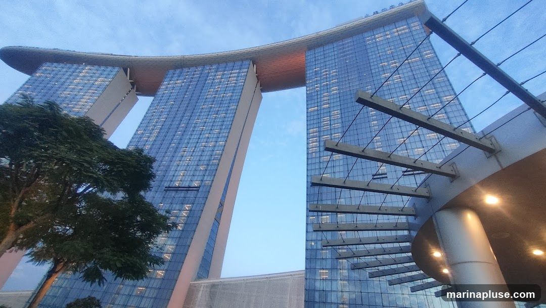 A low-angle shot of the Marina Bay Sands hotel in Singapore. The iconic three towers are visible, connected by the SkyPark. The building's glass facade reflects the blue sky and interior lights. A tree is in the foreground on the left, and a modern architectural canopy with lights is on the right.