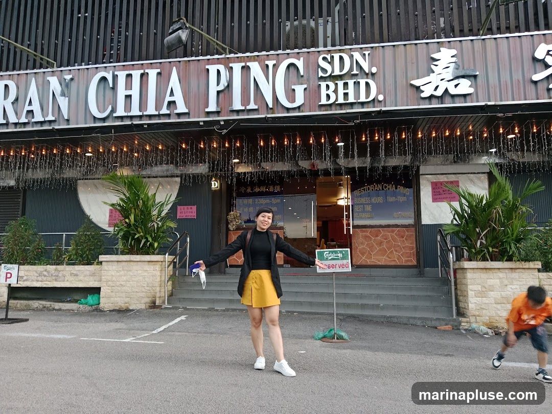 The exterior of a restaurant with a large sign that reads "RAN CHIA PING SDN. BHD." and Chinese characters. There are lights strung across the facade. A woman is standing in front of the entrance, and a child is running in the foreground. A sign for Carlsberg and a "Reserved" sign are visible.