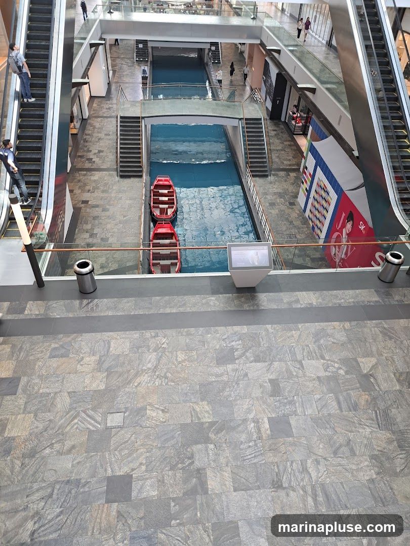 An overhead view of a modern indoor shopping mall featuring a canal with two red boats. Escalators are visible on either side, and people are seen walking on the upper levels and escalators. A promotional display with colorful blocks and an image of a person is on the right side.