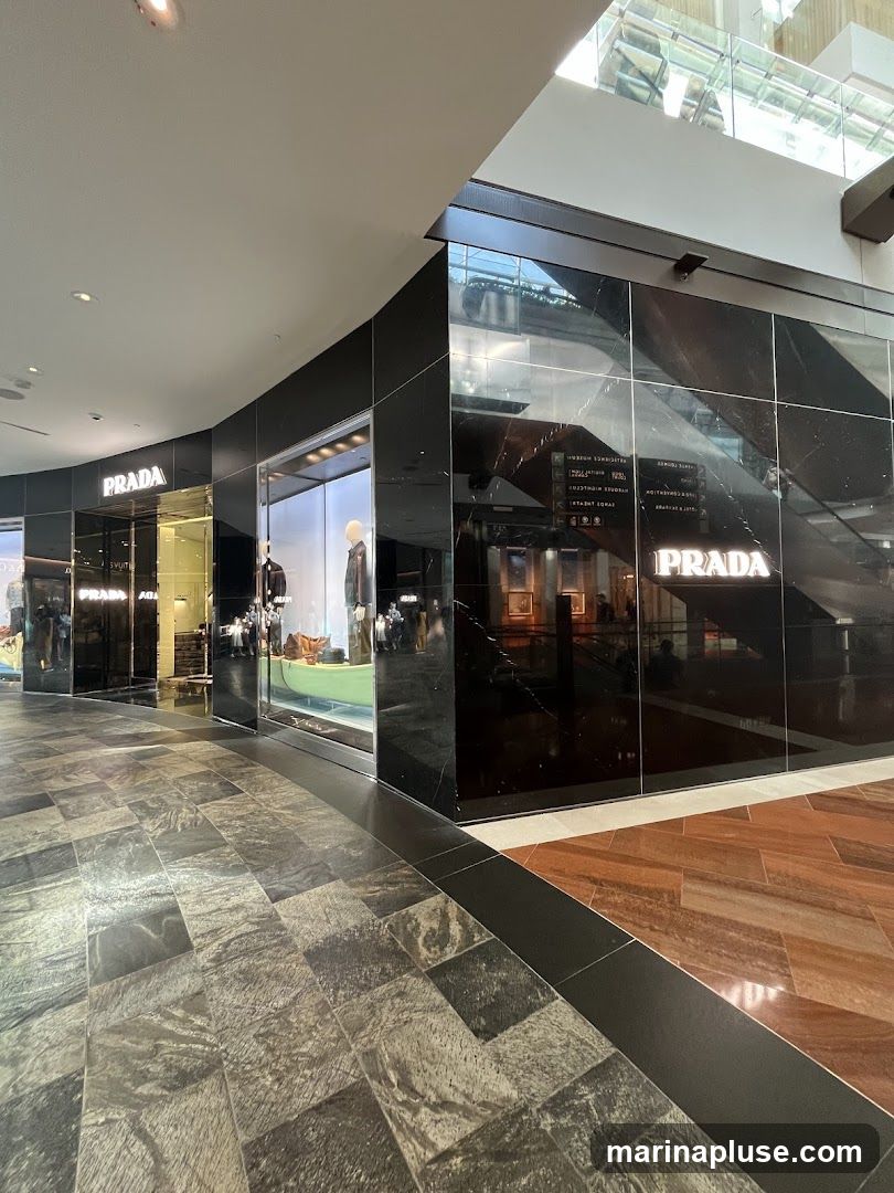 A view of the exterior of a Prada store in a modern shopping mall. The storefront features large glass windows displaying mannequins in fashionable clothing. The facade is made of black marble with the Prada logo illuminated in white. The floor in front of the store is tiled with a dark, patterned stone.