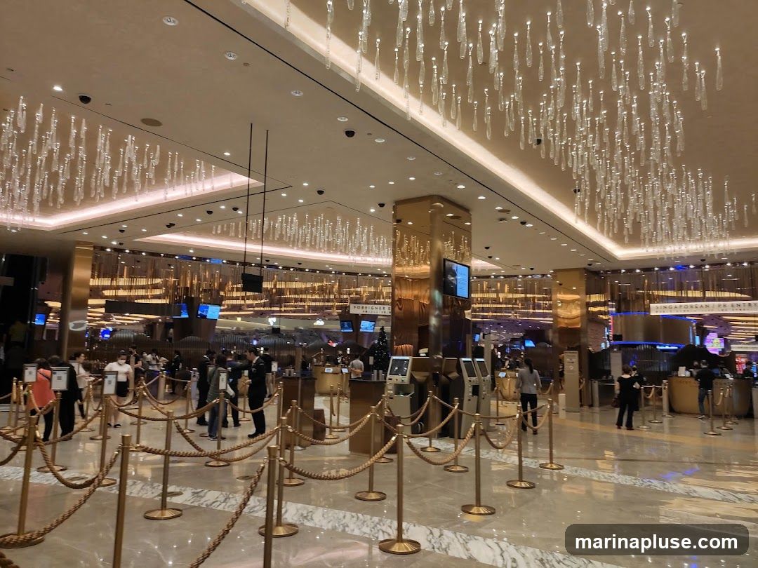 A wide shot of a casino lobby with ornate chandeliers hanging from the ceiling. People are seen queuing behind velvet ropes, and there are various information kiosks and counters visible. Signage indicates separate entry points for "SINGAPOREAN PR ENTRY" and "FOREIGNER ENTRY". The overall ambiance is luxurious and grand.
