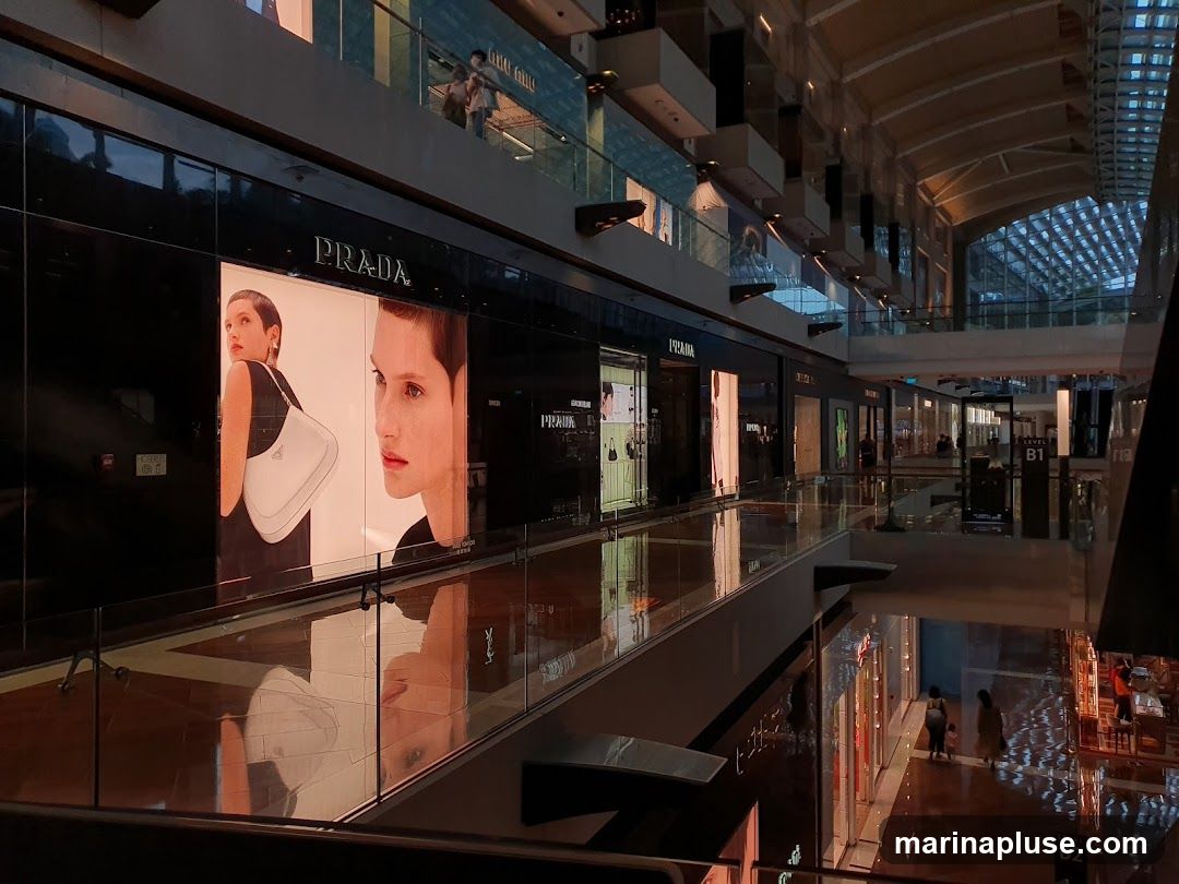 A view of a Prada store and other shops in a modern mall. The Prada storefront features large illuminated displays with images of a woman and a handbag. The mall has multiple levels with glass railings and a distinctive arched ceiling. Reflections of the shops and lights are visible on the polished floor.