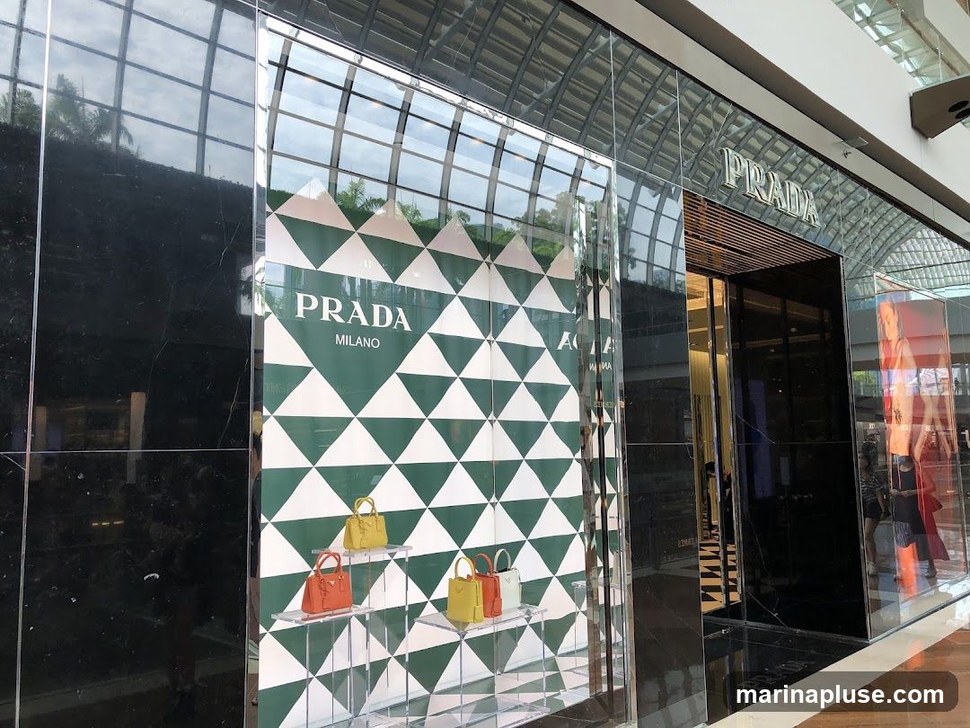 The exterior of a Prada store with its name prominently displayed above the entrance. The storefront features large glass windows showcasing handbags and a geometric green and white pattern. The facade is sleek and modern, with black marble accents and reflective surfaces.