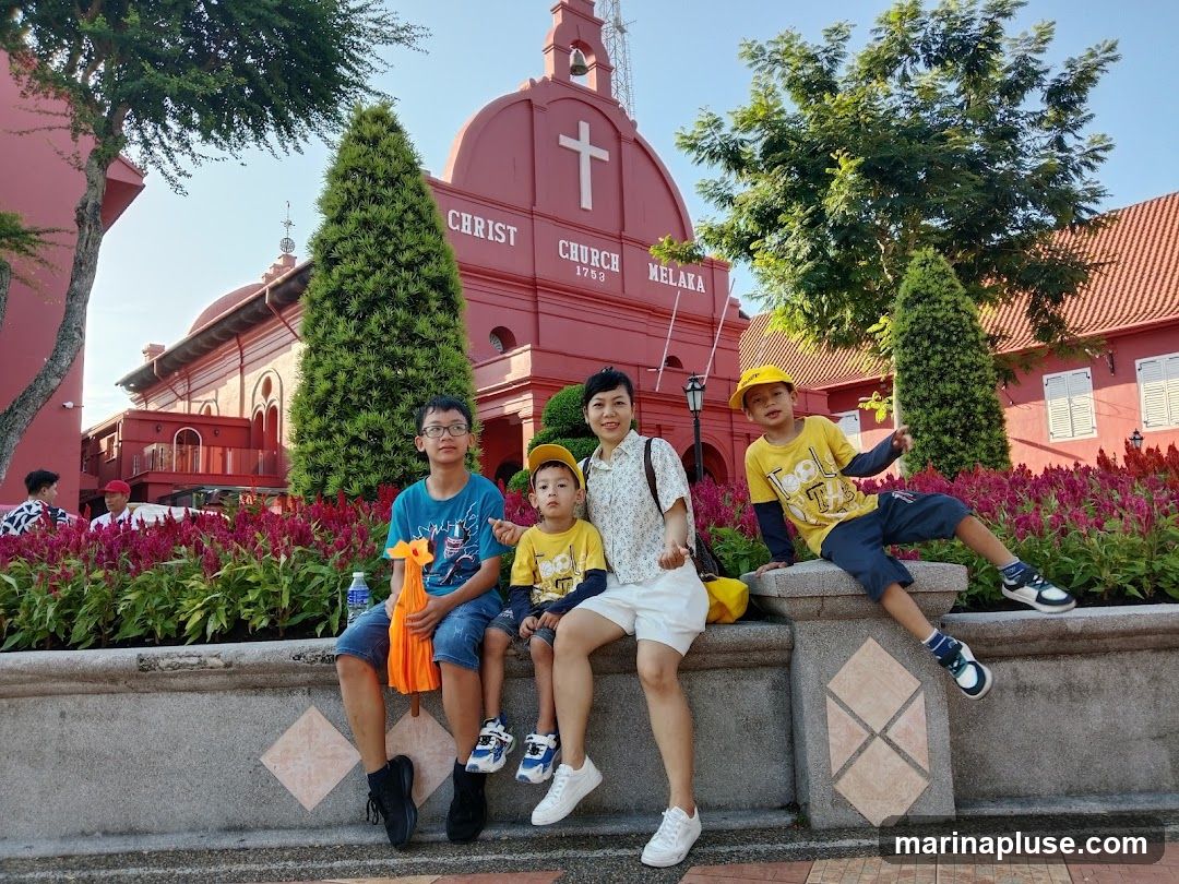 A family poses in front of the red Christ Church Melaka building. The church has a white cross on its facade and the words "CHRIST CHURCH MELAKA 1753" are visible. The building is surrounded by lush greenery and vibrant pink flowers.