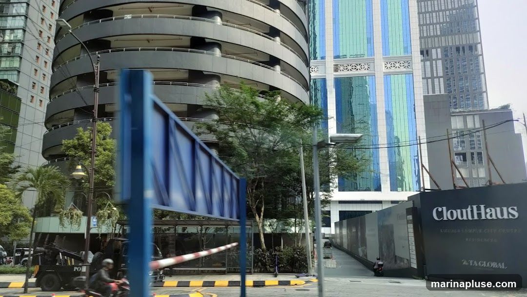 A construction site with a large black hoarding displaying the name "CloutHaus" and "KUALA LUMPUR CITY CENTRE RESIDENCES". Behind the hoarding, modern skyscrapers with glass facades are visible, including a circular parking garage and a building with blue and green vertical stripes. The scene is set on a street with a tow truck and a person on a motorcycle.