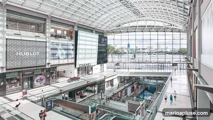 An interior view of a large, modern shopping mall with a glass roof and expansive windows overlooking a waterfront. Multiple levels of shops are visible, including prominent storefronts for HUBLOT and CHANEL. People are seen walking on escalators and walkways. The architecture is sleek and contemporary, with a focus on natural light.