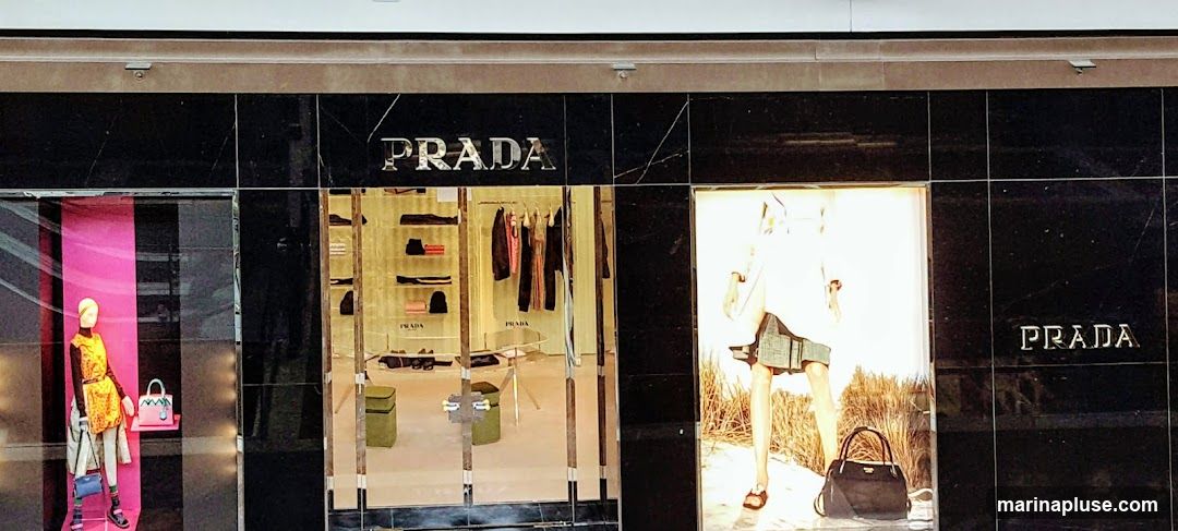 The exterior of a Prada store with large display windows. One window features a mannequin dressed in a yellow and orange patterned outfit with a pink handbag. Another window displays clothing on racks and shelves, with shoes and bags visible. A third window shows a large advertisement with a model wearing a white top and a denim skirt, holding a black handbag. The Prada logo is prominently displayed on the facade above the windows.