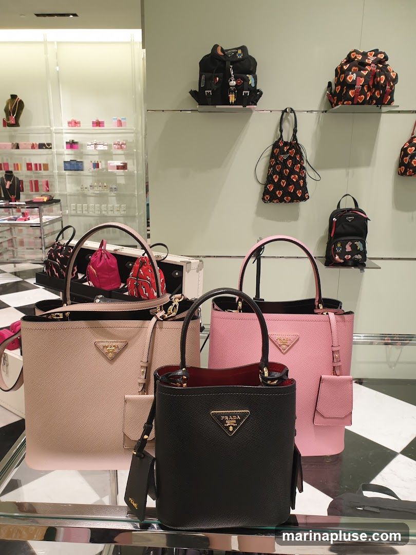 A display of Prada handbags and backpacks in various colors and styles. In the foreground, three handbags are showcased: a beige tote, a black bucket bag, and a pink tote. Behind them, on shelves, are several backpacks, including one black backpack adorned with charms and another with a heart pattern. Smaller bags and accessories are visible on shelves in the background. The display features a black and white checkered floor.