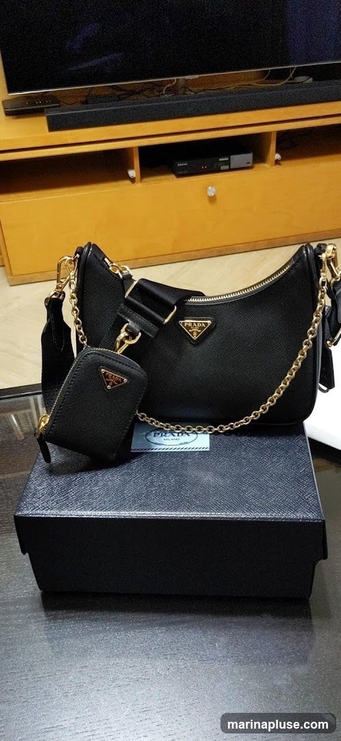 A black Prada handbag with a matching pouch is displayed on top of a black Prada box. The bag features a gold chain strap and a black fabric strap. The Prada logo is visible on both the bag and the pouch. A Prada tag is also visible near the bag. The background shows a wooden cabinet with a television and soundbar.