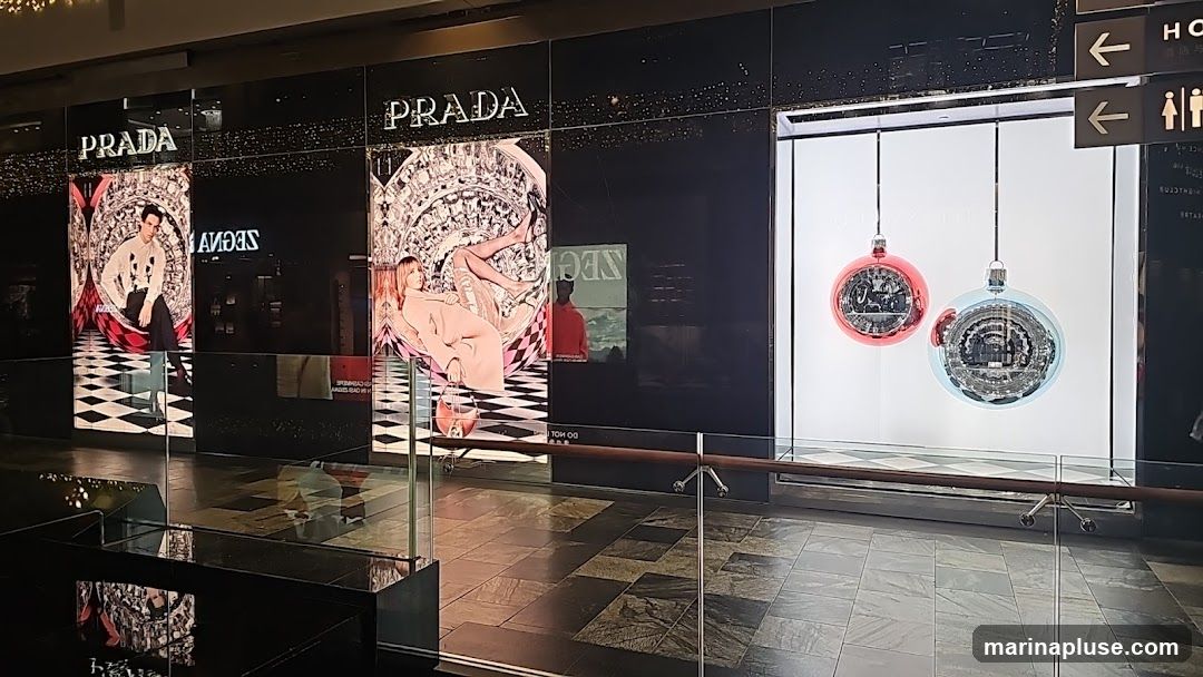 A storefront display for Prada featuring large illuminated panels with fashion imagery and holiday ornaments. The Prada logo is visible above the panels. The display includes images of models and intricate circular designs. Two large, transparent Christmas ornaments, one red and one blue, hang in a separate section of the window. Signage for restrooms is visible to the right.