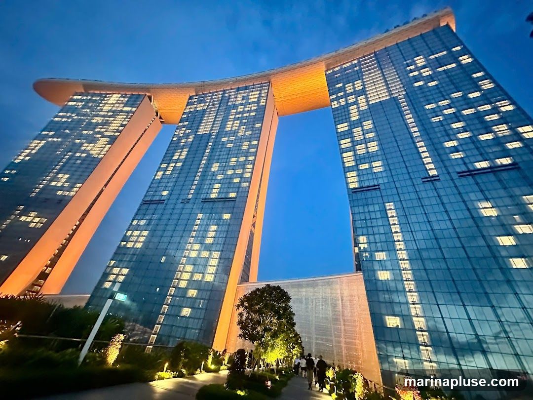 A low-angle shot of the Marina Bay Sands hotel in Singapore at dusk. The three towers are illuminated with warm orange light on their undersides and the windows of the hotel rooms are lit, creating a grid pattern against the blue sky. The iconic SkyPark is visible connecting the tops of the towers. In the foreground, a pathway winds through landscaped gardens with accent lighting. Several people are walking along the path.