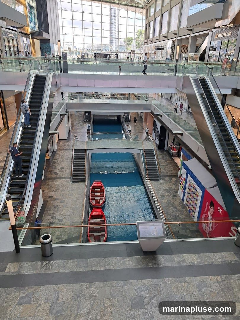 An interior view of a multi-level shopping mall featuring escalators, a canal with gondola-like boats, and various shops. The mall has a large glass ceiling allowing natural light to illuminate the space. People are seen walking on the different levels and riding the escalators. A digital display screen is visible near the canal.