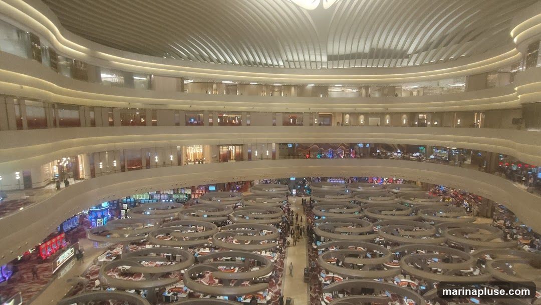 A high-angle view of a multi-level casino interior. The foreground shows rows of circular gaming tables with people seated around them. Above, multiple levels of balconies overlook the casino floor, with various shops and entertainment areas visible. The ceiling features a modern, curved design with integrated lighting.