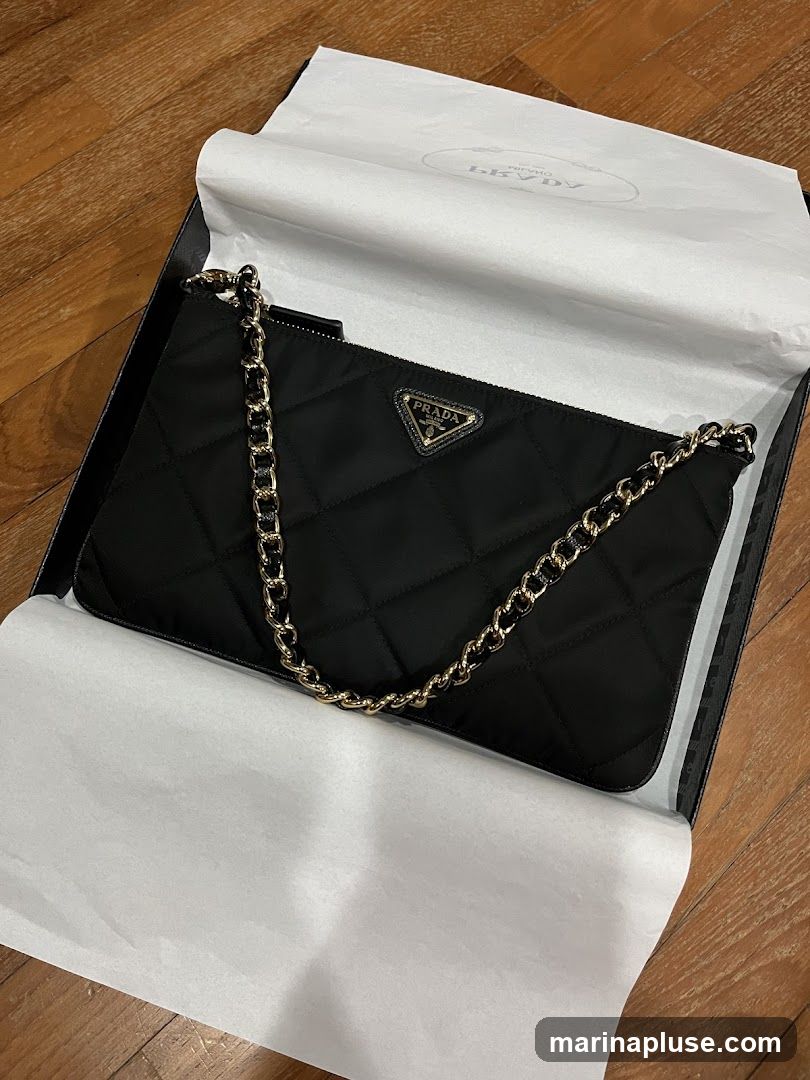 A black quilted Prada clutch bag with a gold chain strap is displayed inside a black box lined with white tissue paper. The Prada logo is visible on the front of the bag. The tissue paper has a repeating Prada logo pattern.