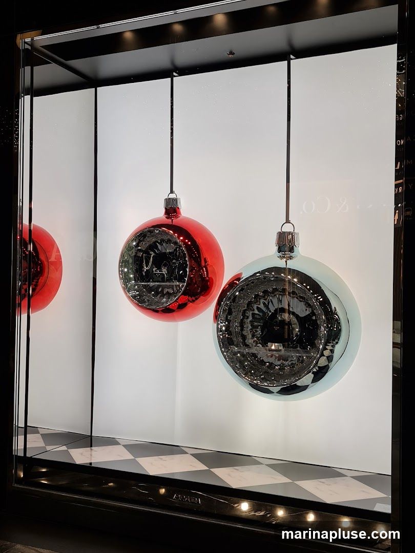 A store window display features large, reflective Christmas ornaments hanging from the ceiling. One ornament is red, and another is a pale blue or silver. The ornaments have a textured, mirrored interior. The background is white and subtly speckled. The floor of the display has a black and white checkered pattern. A faint reflection of text, possibly a brand name, is visible on the glass.