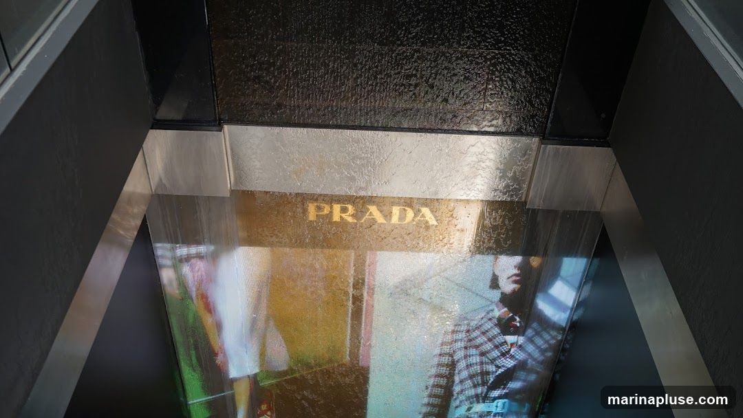 A close-up view of a Prada storefront with the brand name prominently displayed in gold lettering on a metallic surface. Below the name, a digital display shows fashion imagery, including models wearing clothing. The surrounding architecture features dark, textured walls and metallic accents, creating a modern and luxurious aesthetic.