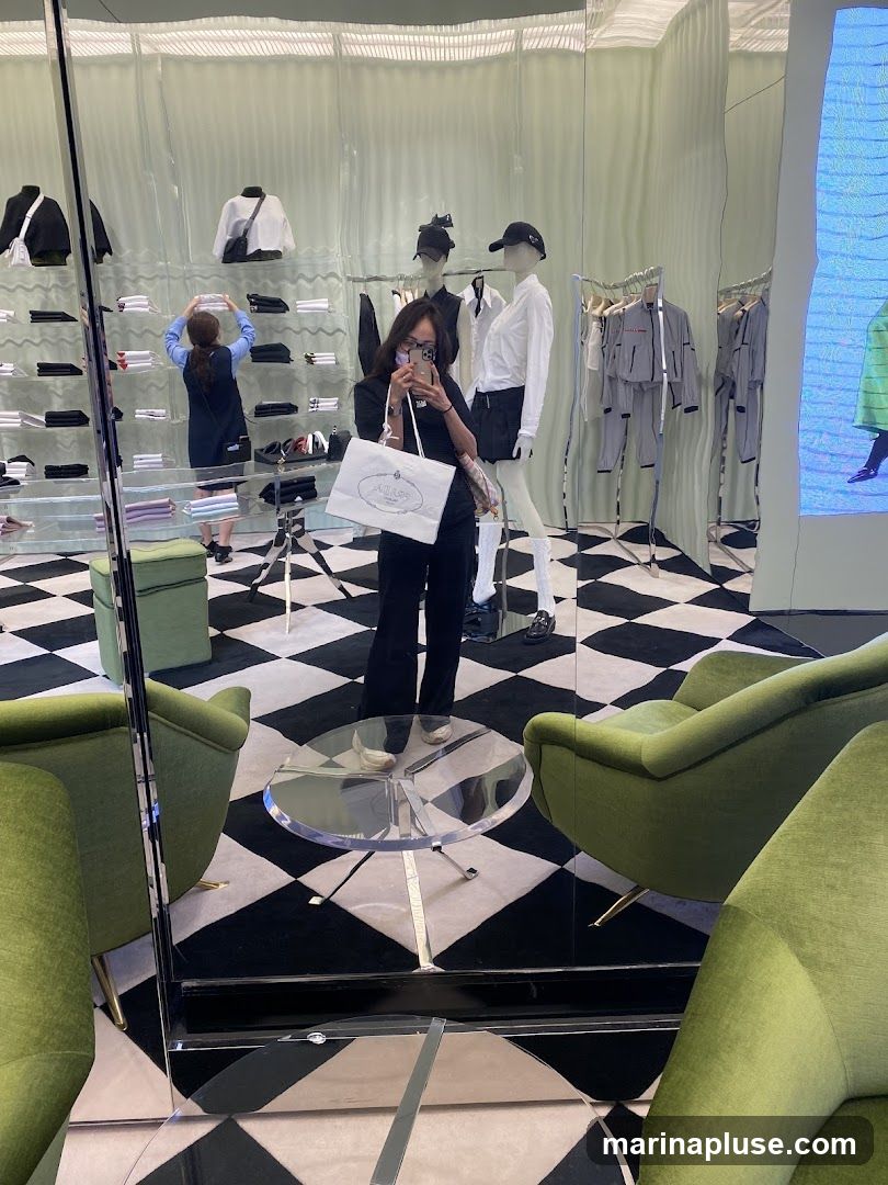 A woman stands in front of a mirror, taking a selfie. She is holding a shopping bag with a logo visible. The store interior is reflected in the mirror, showing clothing on mannequins and shelves, as well as green armchairs and a checkered floor.