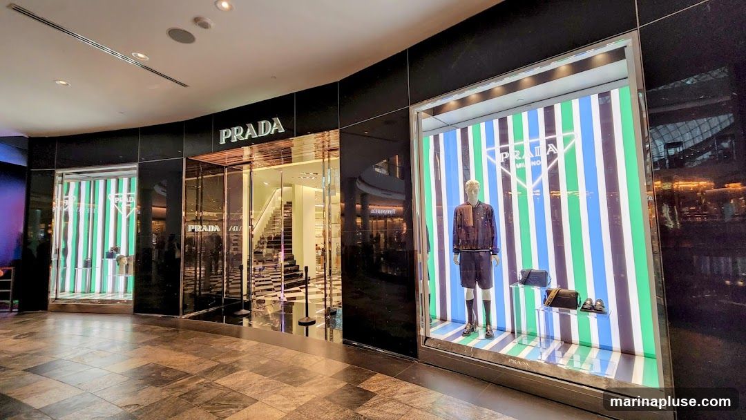 The exterior of a Prada store with a prominent "PRADA" sign above the entrance. The storefront features large display windows showcasing mannequins and merchandise against a backdrop of colorful vertical stripes. The entrance area has a checkered floor and a staircase visible inside.